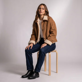 Model seated on a wooden stool in the open Greta jacket showing the cream shearling lining