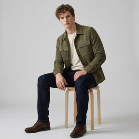Model seated on a wooden stool wearing the olive Dorian overshirt open with dark jeans