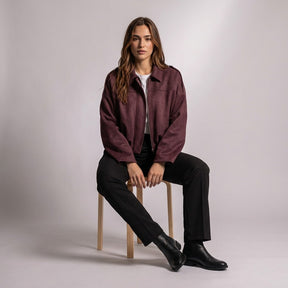 Model seated on a wooden stool wearing the Burgundy Brisa suede bomber